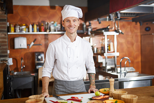 Waist up portrait of handsome chef posing in kitchen and smiling at camera, copy space