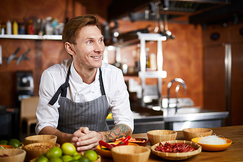 Waist up portrit of handsome chef posing in kitchen at table with spices looking away pensively, copy space