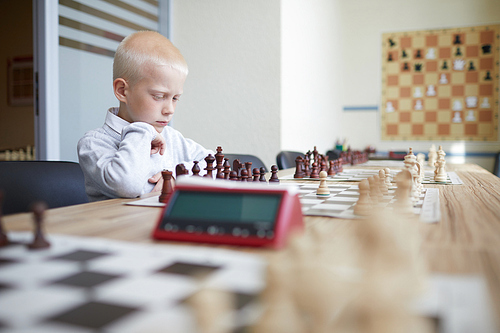 Blonde schoolboy in white shirt playing chess with imaginary opponent in big room of chess club