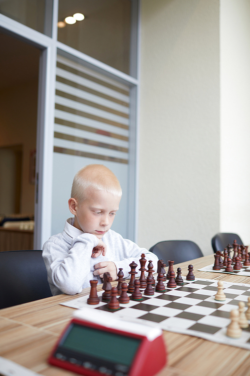 Blonde schoolboy in white shirt looking at board, analyzing it and thinking what chess move to do