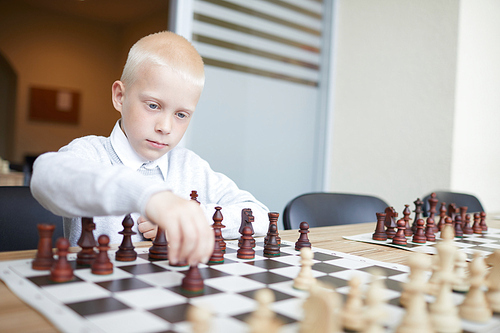 Blonde schoolboy in white shirt making first chess move in game with imaginary opponent