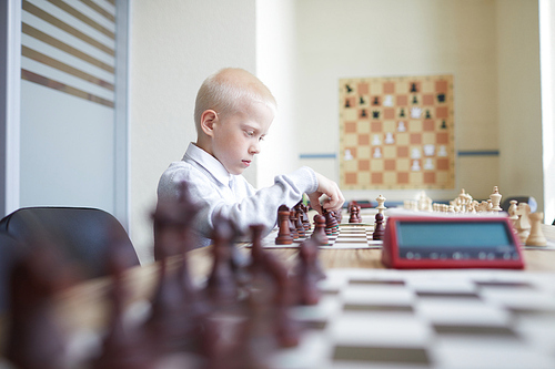 Blonde schoolboy in white shirt playing chess with imaginary opponent and hesitating about next move