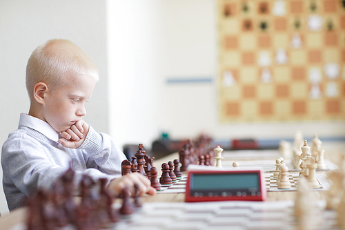 Blonde schoolboy in white shirt seriously thinking about chess game with imaginary opponent