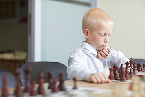 Blonde schoolboy in white shirt attentively studying chessboard and analyzing chess strategy