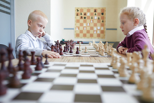 Blonde boy and girl with braided red hair playing chess at table in classroom of chess club