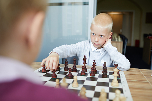 Blonde schoolboy making chess move and looking seriously and confidently into eyes of opponent