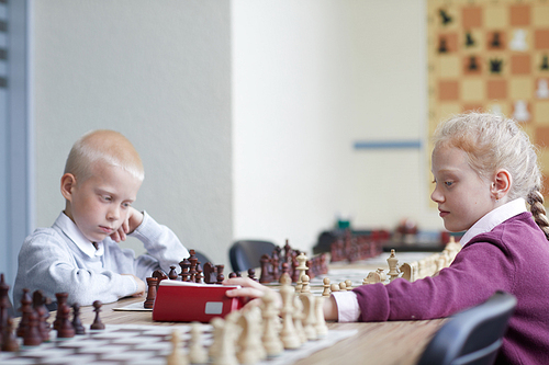 Schoolgirl with red hair pressing chess clock while boy in white shirt thinking carefully about next move