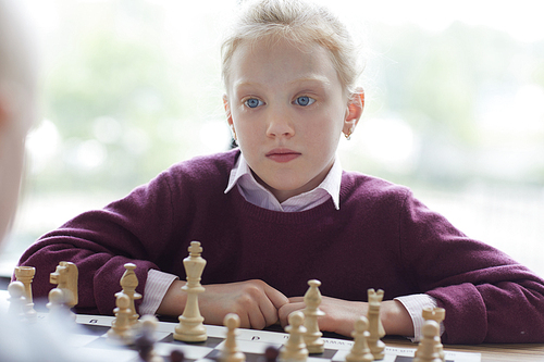 Blue-eyed girl with red hair in purple sweater playing chess and looking attentively at her opponent move