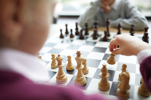 Hand of girl in purple sweater making first chess move in chess game against boy in white shirt