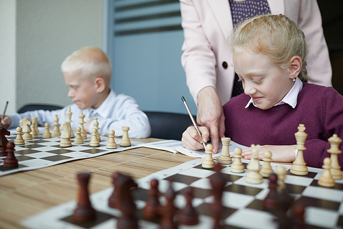 Smiling schoolgirl in purple sweater writing down chess rules while teacher pointing at notebook