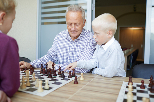 Blonde schoolboy suggesting his grandfather chess move in chess game against his younger sister