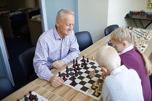 Grandfather in checked shirt playing chess against his granddaughter with red hair and his blonde grandson