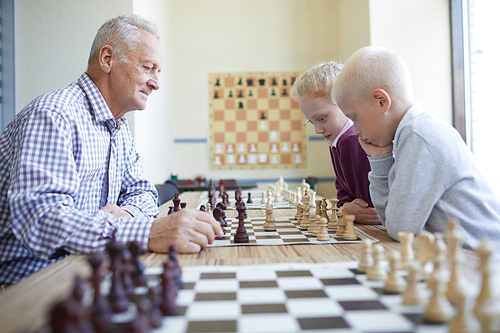 Old man in checked shirt playing chess with two focused schoolchildren studying at chess club