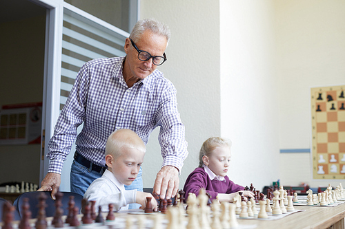 Aged male teacher with glasses helping two schoolchildren with learning chess combinations during chess class