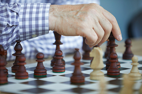 Tanned wrinkled hand of old man in checked purple and white shirt making chess move with dark figures