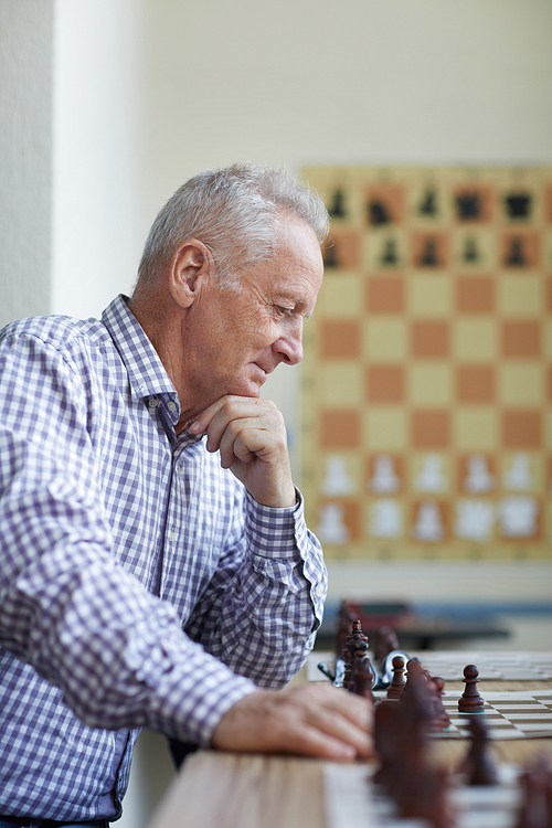 Old man in checked shirt with silver hair looking with smile at chess board and analyzing interesting chess game