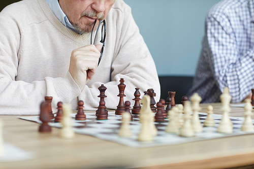 Aged focused experienced chess player with grey beard biting glasses while analyzing chess game