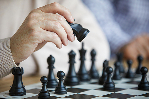 Wrinkled hand of old chess player in beige sweater moving black chess figure along chess board