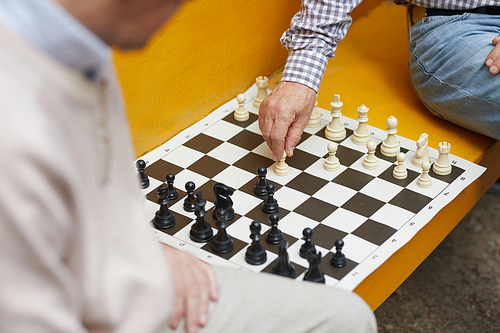 Wrinkled hand of old chess player moving white figure during playing chess on yellow bench