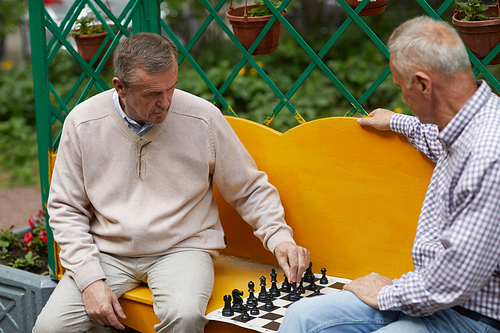 Two aged brothers with silver hair sitting outside on yellow bench and playing chess at backyard