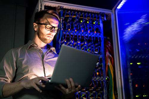 Serious busy young IT engineer in glasses standing against network server equipment with glowing sensors in dark room and using laptop while monitoring network systems