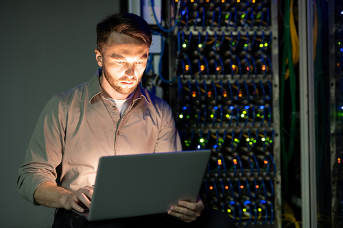 Concentrated young bearded server manager in shirt working with laptop in dark room of data center