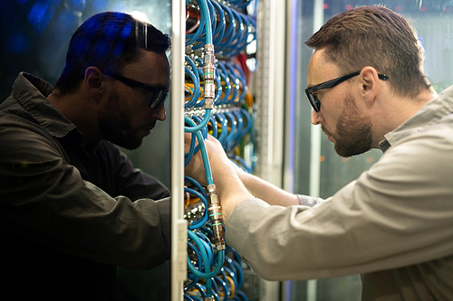 Side view of concentrated young server repairman in glasses standing in server room and connecting cables to supercomputer