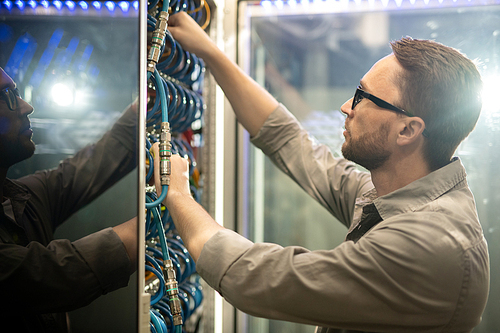 Serious skilled young data center engineer in glasses standing at mainframe and examining cables while doing checkup