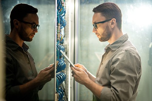 Serious pensive young IT engineer with beard standing by server cabinet and using tablet while setting up server network