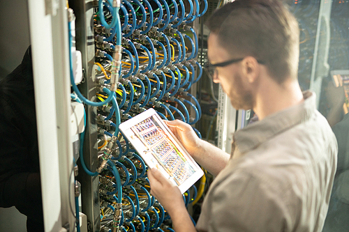 Busy server engineer standing by open cabinet of network server and using modern device while analyzing connections
