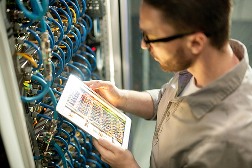 Close-up of concentrated man in shirt standing by mainframe and using digital tablet to check connectivity in datacenter room