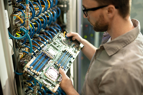 Close-up of serious bearded IT support specialist standing by cabinet of mainframe and examining motherboard of server