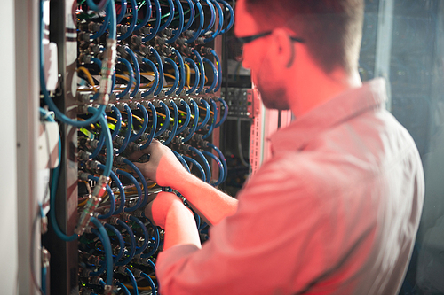 Rear view of skilled man standing by cabinet of server racks and examining fiber cables while repairing server