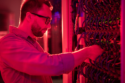 Serious concentrated bearded network technician in glasses standing at cabinet of mainframe and fixing supercomputer, red light