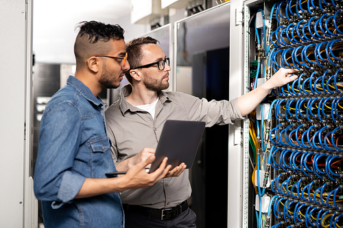 Serious thoughtful young multi-ethnic computer support specialists in casual clothing standing at server racks and using laptop while analyzing network problem