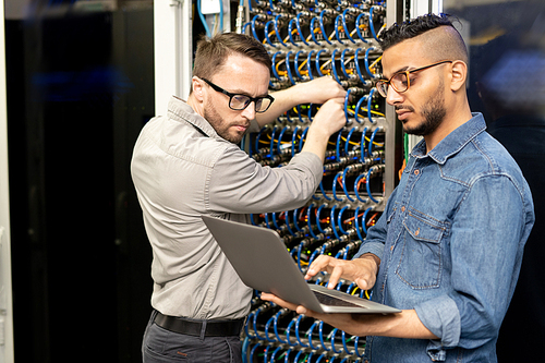 Serious young multi-ethnic IT engineers in casual shirts standing in database center and setting up computer cluster while examining information on laptop