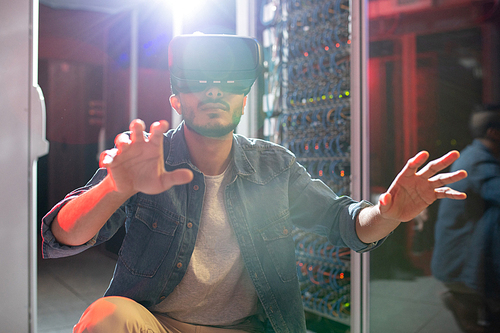 Serious concentrated young Arabian IT engineer with beard crouching on floor of data center and gesturing hands while studying server systems using VR goggles