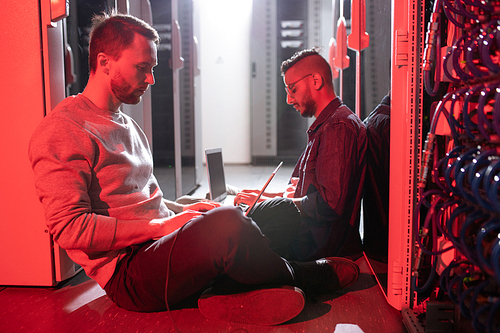 Concentrated young multi-ethnic engineers sitting on floor in server room with red light and working with laptops