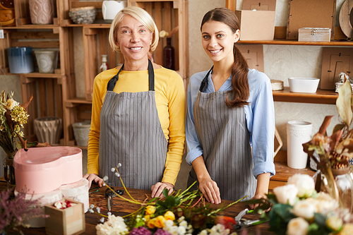 Waist up portrait of two female florists smiling at camera while arranging flowers and posing in flower shop