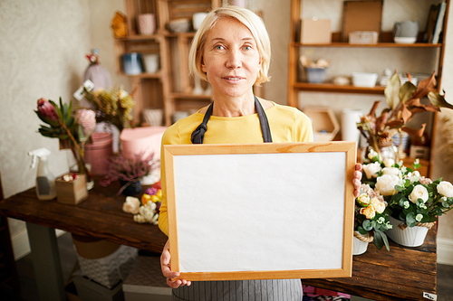 Waist up portrait of mature female florist holding whiteboard and   while working in flower shop, copy space