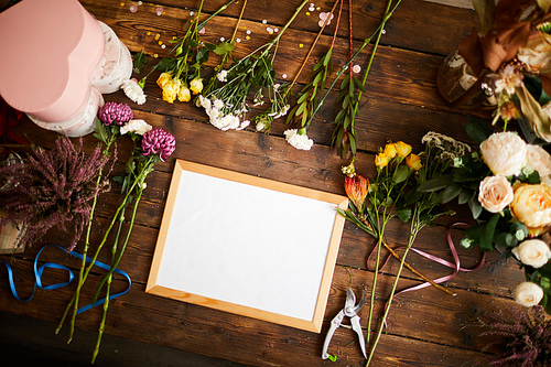 Top view of blank whiteboard on wooden table in flower shop, surrounded with beautiful blooms and creative supplies