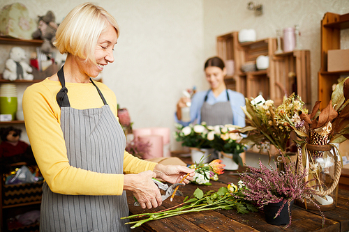 Side view portrait of smiling female florist cutting flower stems while arranging bouquets in flower shop, copy space