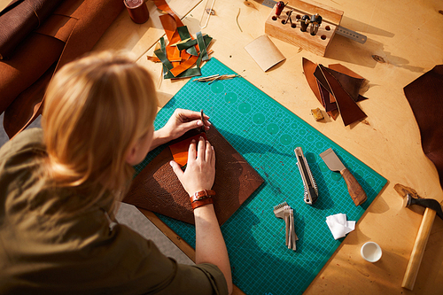 Above view portrait of female artisan making leather bag in leatherworking atelier lit by sunlight, copy space