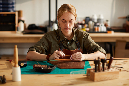 Portrait of young woman working with leather while making handcrafted belt in shop lit by sunlight