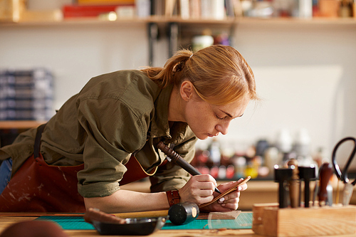 Portrait of woman artisan working with leather while making handcrafted belt in shop lit by sunlight