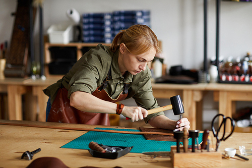 Portrait of woman artisan making leather belt in shop lit by sunlight, copy space