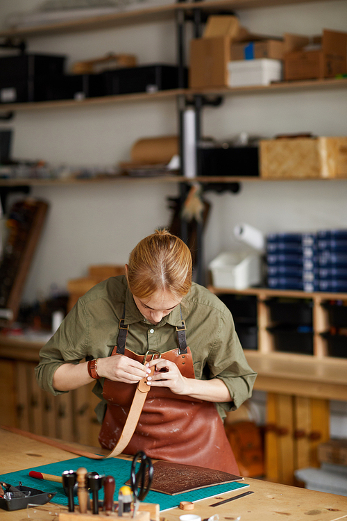 Waist up portrait of woman artisan making leather belt in shop lit by sunlight, copy space