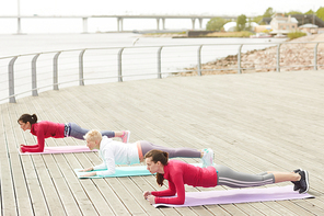 Full length portrait of three contemporary women working out outdoors at dawn standing in plank position on wooden pier, copy space