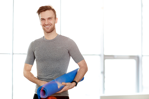Content confident handsome male yoga instructor with exercise mat standing against window and smiling at camera