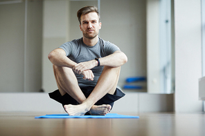 Serious pensive young man sitting on exercise mat and embracing knees while waiting for yoga class, he 